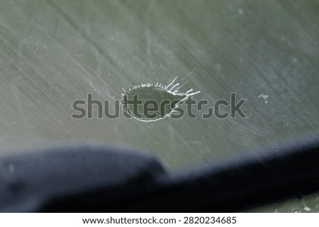 A teardrop ring marks the glass while a dark wiper edge crosses the frame. Scratches and soft green light complete the scene. The quiet composition suggests travel in gentle rain.