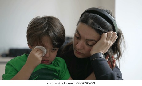 Tearful child holding cotton into hurt eye with patient but tired mother waiting for emergency service at hospital - Powered by Shutterstock