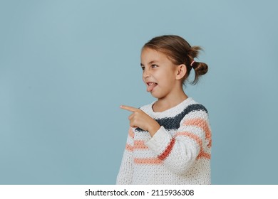 Teasing nasty little girl in striped sweater points her finger and sticks out tongue over blue background. Side view.