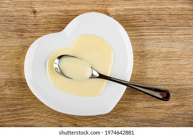 Teaspoon with condensed milk in white glass saucer in heart shape on brown wooden table. Top view