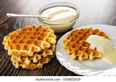 Teaspoon in transparent bowl with condensed milk, stack of waffles, white plate with biscuit waffle poured condensed milk on wooden table
