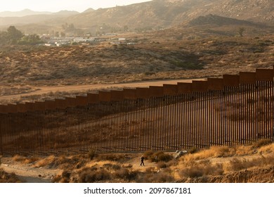 Tecate, Baja California, Mexico - September 14, 2021: Late Afternoon Sun Shines On The USA Mexico Border Wall People Walk In Front Of It.