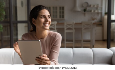Tech and dreams. Happy dreamy latin woman sit on couch work on tablet online look at distance think ponder on good result. Calm young female use pad at home take break create idea imagine. Copy space