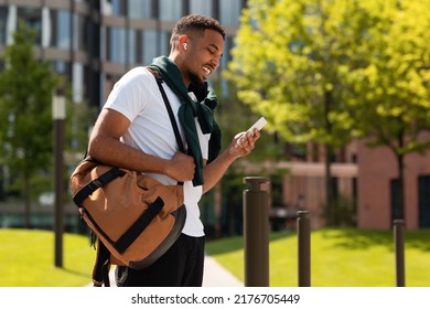 Tech gadgets for entertainment. Happy african american stylish guy walking and using smartphone, listening music in earpods, standing outdoors in ubran park area