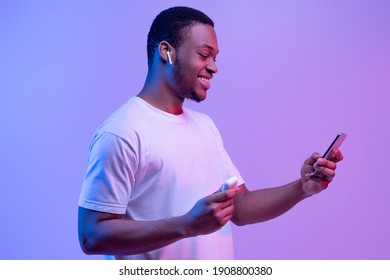 Tech Gadgets For Entertainment. Happy Black Guy Using Smartphone And Earpods For Listening Music, Holding Earphones Case In Hand, Cheerful African Man Standing In Neon Light Over Purple Background