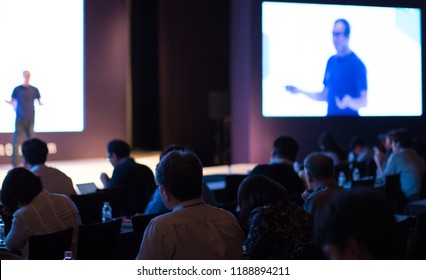 Tech Presenter On Stage Giving Presentation Speech At Conference Seminar. Innovation And Growth Speaker Giving Lecture At Meeting. Audience Watching Presenter. White Screen Background