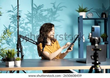 Tech specialist presenting tablet in entry level low budget price range to interested viewers, filming with smartphone on tripod. Woman reviewing digital device, recording with mobile phone