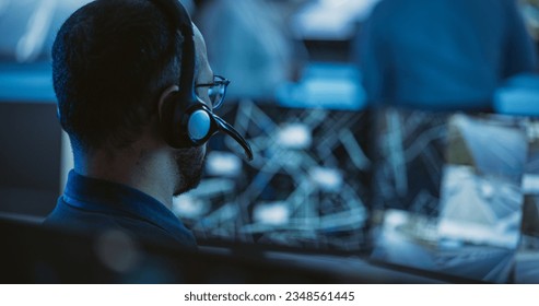 Technical Customer Support Specialist is Talking on a Headset while Working on a Computer in a Call Center Control Room Filled Display Screens and Data Servers. Footage from the Back - Powered by Shutterstock