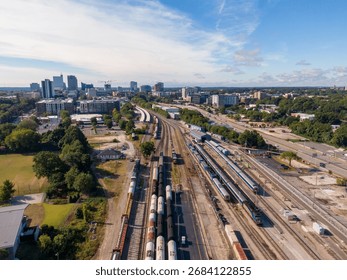 A technical high-angle drone perspective documenting multiple passenger and freight trains near the downtown Raleigh, North Carolina skyline. The shot captures a consist of Amtrak Piedmont and Carolinian passenger cars, which are part of the state-sponsored 