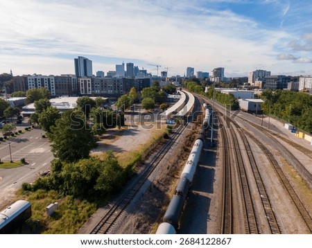 A technical high-angle drone perspective documenting multiple passenger and freight trains near the downtown Raleigh, North Carolina skyline. The shot captures a consist of Amtrak Piedmont and Carolinian passenger cars, which are part of the state-sponsored 