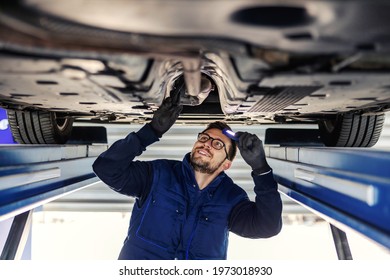 Technical inspection of the car. Car service in the workshop. A man in a blue uniform stands under a car in the garage and checks the car’s axles. It illuminates the chassis with a flashlight - Powered by Shutterstock