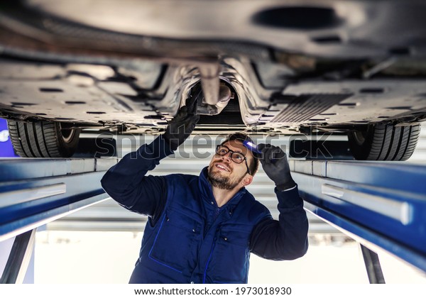 Technical inspection of the car. Car service\
in the workshop. A man in a blue uniform stands under a car in the\
garage and checks the car’s axles. It illuminates the chassis with\
a flashlight