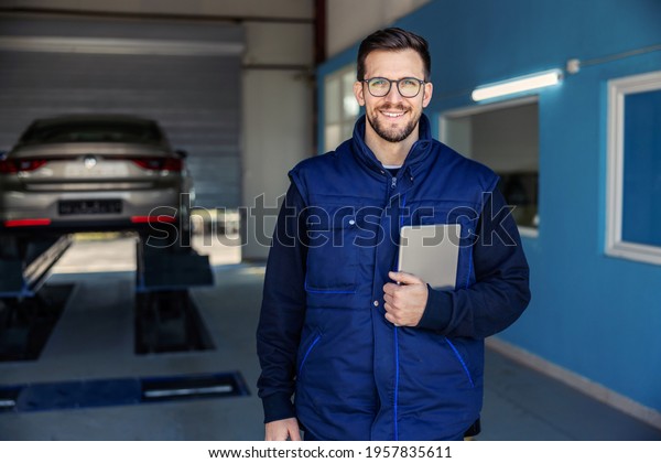 Technical inspection of cars and tablets. A man\
in uniform holds a tablet in his hands in a workshop in front of a\
car on a hydraulic elevator. Portrait of a man at work in a\
workshop, car service