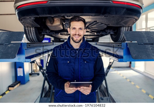 Technical inspection of cars and tablets. Man in\
uniform holds a tablet in his hands in the workshop canal in front\
of a car on a hydraulic elevator Portrait of a man at work in a\
workshop Car service