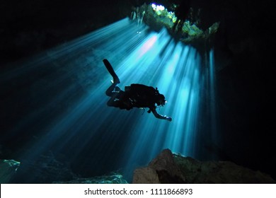 Technical scuba diver exploring underwater cenote. Scuba diver in the sun light from the water surface. Underwater dark cave and diver.