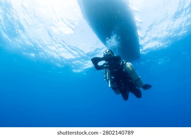 A technical sidemount diver ascends through clear blue ocean water toward a boat hull. Sunlight penetrates the surface, creating bright patterns above the diver. The diver wears full gear.