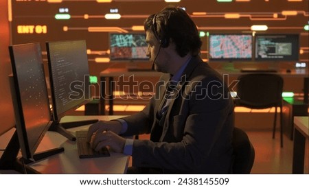 Technical support in control room. Man IT specialist works on computer in headset at the office, big digital screen shows infrastructure data.