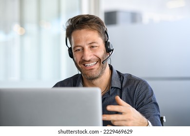 Technical support operator working with headset in office. Smiling handsome man working as call centre operator, speaking to customer. Happy businessman working remotely while doing video conference. - Shutterstock ID 2229469629
