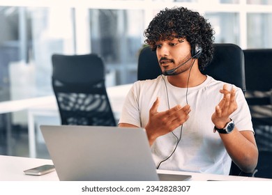 Technical support operator working with headset in office. Smiling handsome man working as call centre operator, speaking to customer. Happy businessman working remotely while doing video conference.