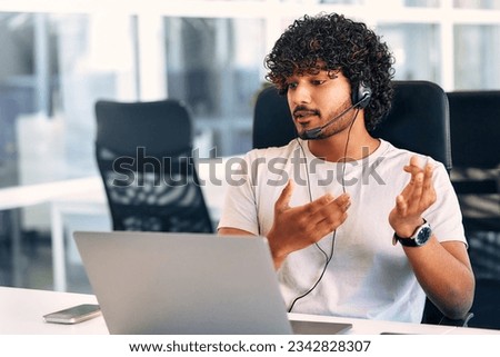 Technical support operator working with headset in office. Smiling handsome man working as call centre operator, speaking to customer. Happy businessman working remotely while doing video conference.