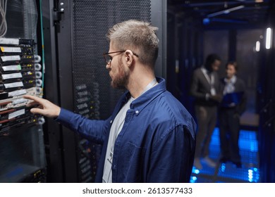 Technician in blue shirt configuring server rack in data center with colleagues in background. LED lights illuminating server room creating modern technological ambiance