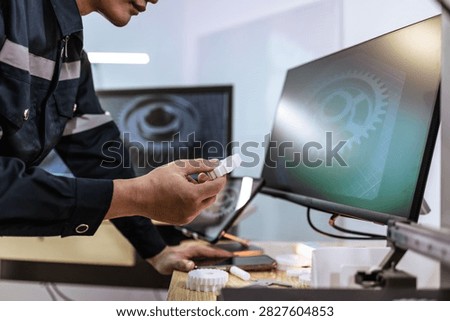 Technician checking a small 3d printed part beside computer screens in a workshop with machine design on display.