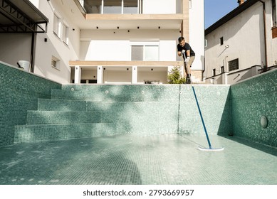 Technician cleans the empty pool by scrubbing the walls and tiles. The pool is being prepared for the upcoming season. The sunlight highlights the geometric design.