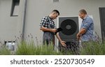 A technician consults with a homeowner about a heat pump, using a tablet for demonstration beside the unit.