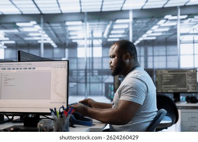 Technician at desk writing code and doing checkup on server room providing computing resources for different workloads. African american man investigating data center mainframes using computer