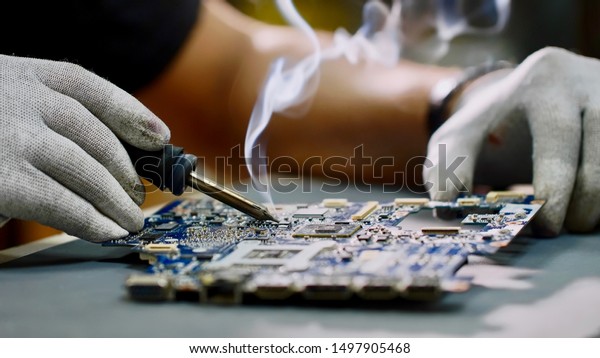 Technician engineer in workshop. Repairman\
in gloves is soldering circuit board of electronic device on the\
table, hands close up. He takes tin with a soldering iron and puts\
it on microcircuit.