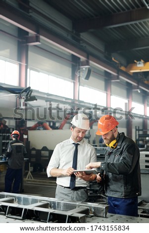 Technician in formalwear using tablet while engineering industrial machine with factory worker in workshop