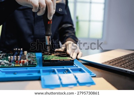Technician in gloves repairs a computer hard drive with a screwdriver at a workbench beside a laptop, showing electronics maintenance, data recovery, and hardware service.