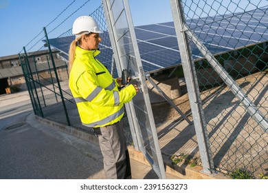 Technician in high visibility jacket on a gate with walkie-talkie at solar farm. Alternative energy ecological concept image. - Powered by Shutterstock