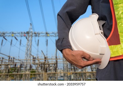Technician holding white helmet on blurry power substation background.