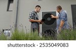 A technician and homeowner discuss heat pump maintenance, referencing a tablet near the unit.