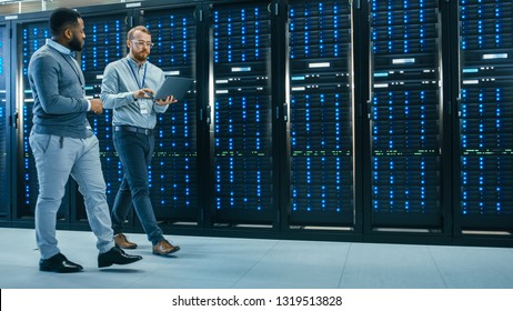 IT Technician With A Laptop Computer And Black Male Engineer Colleague Are Talking In Data Center While Walking Next To Server Racks. Running Diagnostics Or Doing Maintenance Work.