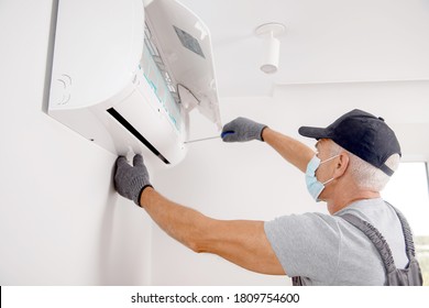 Technician man in medical mask worker repairing and installs air conditioner on white wall.