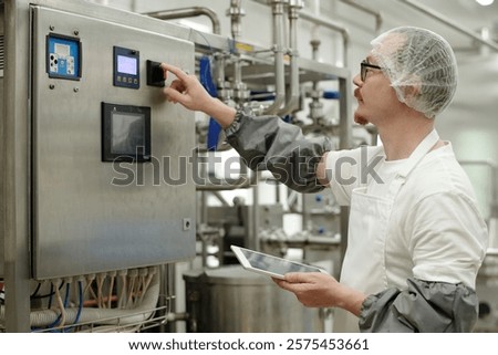 Technician managing electronic control panel in laboratory environment while holding tablet. Wearing safety gear and interacting with advanced technology