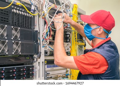 A Technician In A Medical Mask Switches Wires In The Server Room. A Man In A Red Cap Works In A Datacenter. A Technical Employee Maintains Computer Equipment