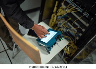 A technician operates a handheld device in a data center. The photo captures a close-up of the technician’s hand using a touchscreen for network analysis, surrounded by networking cables and equipment.