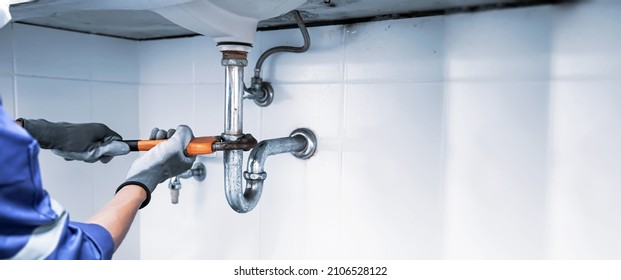 Technician plumber using a wrench to repair a water pipe under the sink. Concept of maintenance, fix water plumbing leaks, replace the kitchen sink drain, cleaning clogged pipes is dirty or rusty. - Shutterstock ID 2106528122