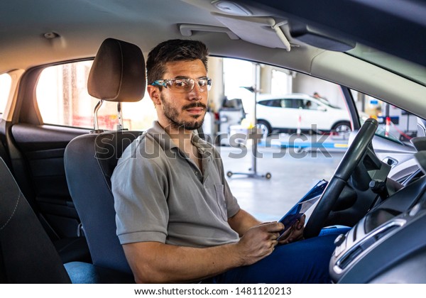 Technician with safety glasses sitting in a\
car seat looking at camera inside a car checking a list during a\
vehicle inspection