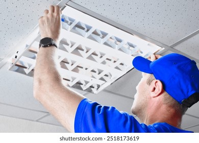 A technician is securing and adjusting a fluorescent light panel in the ceiling of a commercial office, wearing blue work clothes, and ensuring everything is in place for optimal lighting. - Powered by Shutterstock