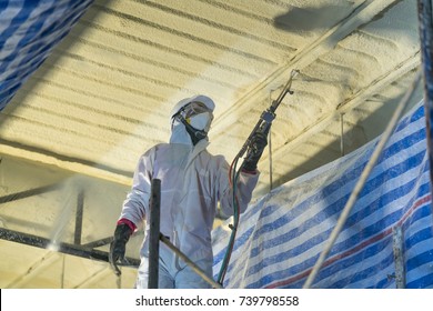 Technician spraying foam insulation using Plural Component Gun for polyurethane foam - Repair tool in the white protect suit applies a construction foam from the gun to the roof of a warehouse. 