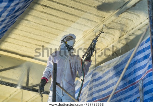 Technician spraying foam insulation using Plural\
Component Gun for polyurethane foam - Repair tool in the white\
protect suit applies a construction foam from the gun to the roof\
of a warehouse. 