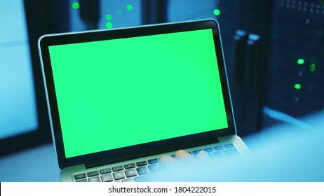 IT technician using a modern laptop in server cabinet, running diagnostics on mock-up greenscreen device, working in advanced high tech data center. Close-up.