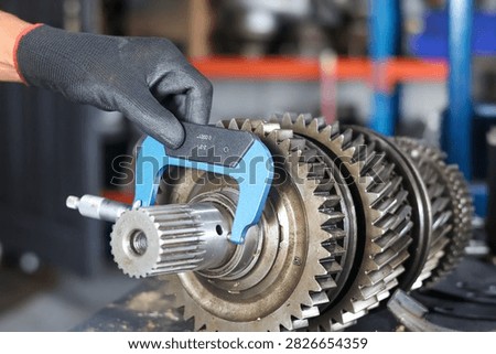 Technician using a precision micrometer tool to measure the shaft dimensions of a manual transmission gear cluster in a workshop.