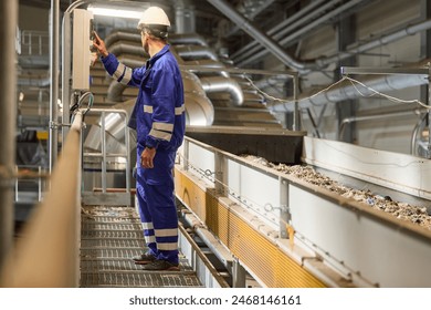 A technician wearing blue overalls and a white helmet operates machinery at an industrial recycling facility where he manages the sorting and processing of refuse-derived fuel (RDF) on a conveyor belt - Powered by Shutterstock