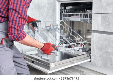 Technician or worker in uniform installs dishwasher into the kitchen furniture. Repairman wear worker suit repairing maintenance of dishwasher. Master in protective gloves fix dishwasher.