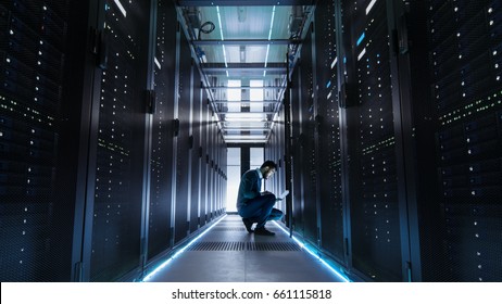 IT Technician Works On Laptop In Big Data Center Full Of Rack Servers. He Runs Diagnostics And Maintenance.